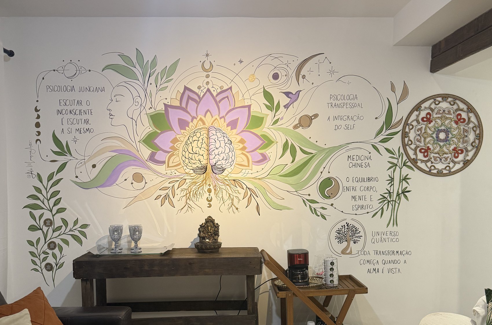Mural pintado na parede do consultório: um cérebro brotando de uma flor de lótus com raízes, cercado pelos dizeres “psicologia junguiana — escutar o inconsciente é escutar a si mesmo”, “psicologia transpessoal — a integração do Self”, “medicina chinesa — o equilíbrio entre corpo, mente e espírito” e “Universoo Quântico — toda transformação começa quando a alma é vista”.